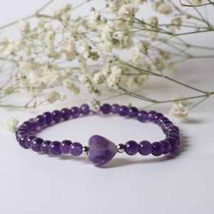 May include: A purple amethyst heart charm bracelet with silver beads. The bracelet is on a white background with white flowers.