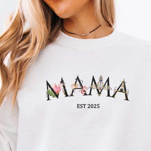 Peut inclure: Sweat-shirt blanc avec le mot "MAMA" en lettres noires avec des accents floraux. Le texte "EST 2025" est sous le mot "MAMA".