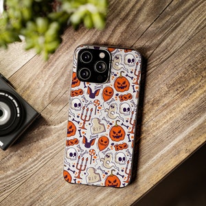 Peut inclure: Une coque de téléphone blanche avec un motif sur le thème d'Halloween, avec des citrouilles orange, des crânes, des fantômes, des chauves-souris et des os. L'étui présente des accents violets et le texte "RIP" sur une pierre tombale. La coque est sur une surface en bois.