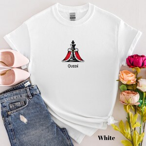 May include: White t-shirt featuring a graphic of a queen chess piece with a red and black design, and the word "QUEEN" below it. The shirt is displayed with blue jeans, pink shoes, and flowers.