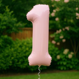 May include: A large, pink number 1 balloon, ideal for a first birthday. The balloon has a shiny surface and is inflated. A white ribbon is attached to the bottom. The background is a blurred green lawn and foliage.