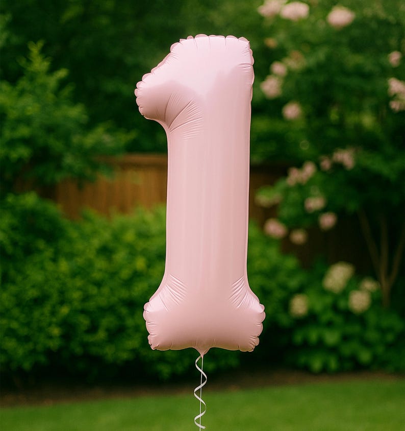May include: A large, pink number 1 balloon, ideal for a first birthday. The balloon has a shiny surface and is inflated. A white ribbon is attached to the bottom. The background is a blurred green lawn and foliage.
