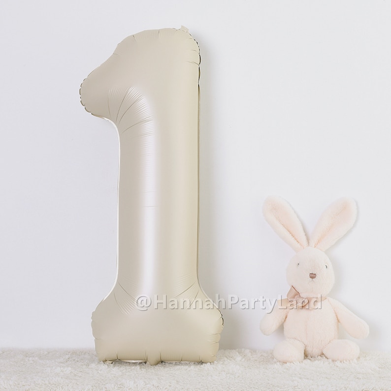 May include: A large, beige, foil number one balloon with a white plush bunny rabbit toy in front of it. The balloon is on a white fluffy surface.