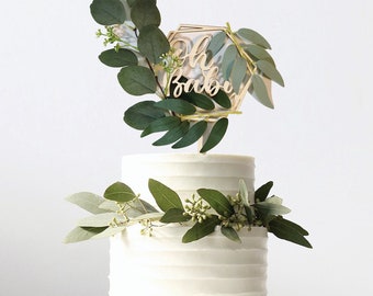May include: A two-tiered white cake decorated with greenery and a wooden cake topper that says "Oh Baby" in a geometric shape.