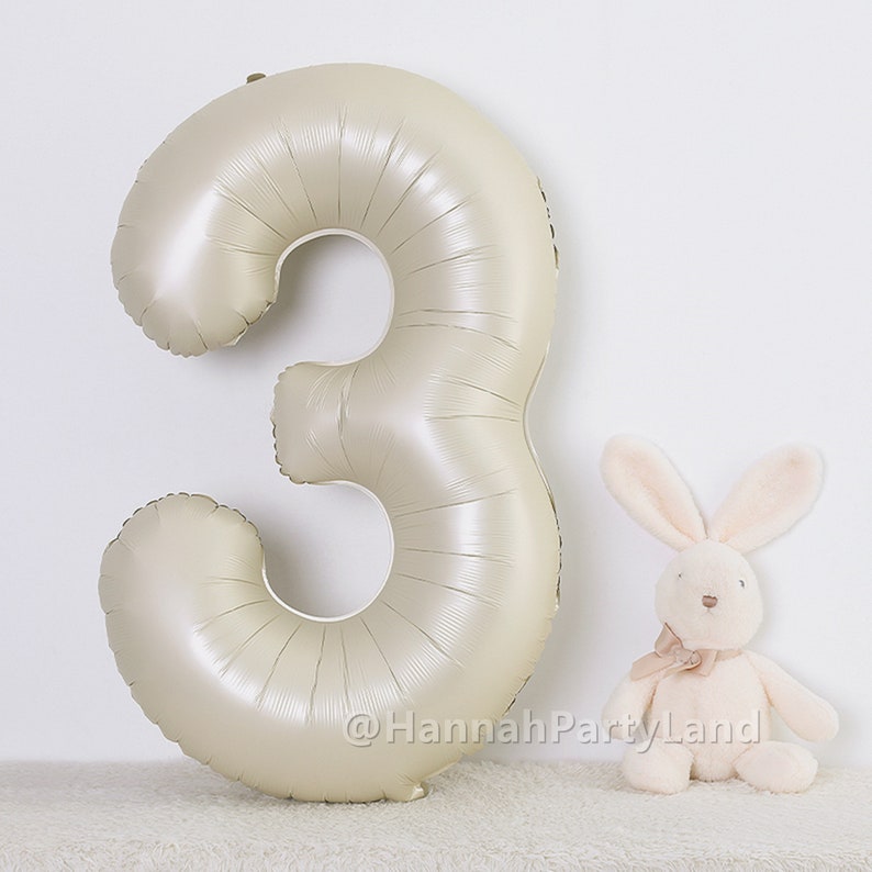 May include: A large, metallic gold number three balloon. The balloon is inflated and has a shiny finish. A small, white plush bunny is sitting in front of the balloon.