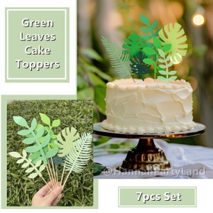 May include: Green leaf cake toppers, a set of 7 paper leaves on sticks, ideal for decorating a cake. The leaves are in various shades of green and feature different leaf shapes, including monstera leaves and fern leaves. The text "7pcs Set" is visible in the image.