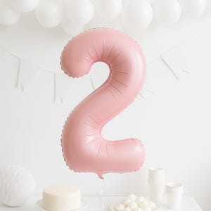 May include: A large, pink number 2 balloon dominates the image, perfect for a second birthday. White balloons, a small cake, and paper cups are also visible, creating a celebratory atmosphere. White pennant flags hang in the background.