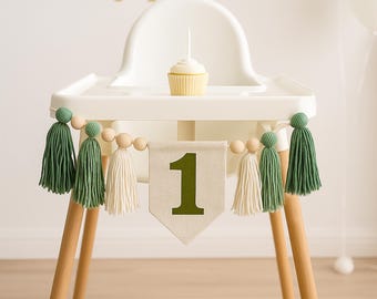 May include: A white high chair with a green and white tassel garland with wooden beads. The garland has a banner with the number "1" on it. A small cupcake is on the tray of the high chair.