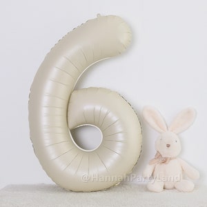 May include: A large, cream-colored, foil number six balloon. The balloon is inflated and standing upright on a white surface. A small, cream-colored, plush bunny is sitting to the right of the balloon.
