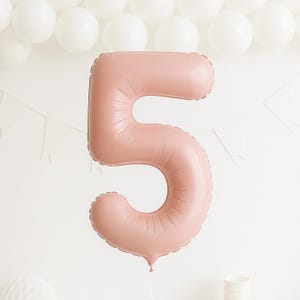 May include: A large, dusty rose number five balloon, ideal for a birthday. The inflated balloon hangs against a white backdrop, with white balloons and a banner in the background. The balloon is in the shape of the number 5.