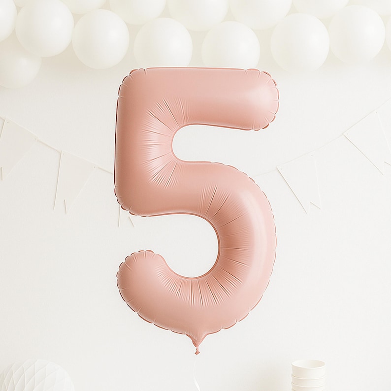 May include: A large, dusty rose number five balloon, ideal for a birthday. The inflated balloon hangs against a white backdrop, with white balloons and a banner in the background. The balloon is in the shape of the number 5.