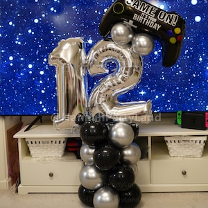 May include: Silver number balloons spelling out "12" with a black balloon shaped like a video game controller with the text "Game On! It's Your Birthday" on it. The balloons are on a white table with a white wicker basket.