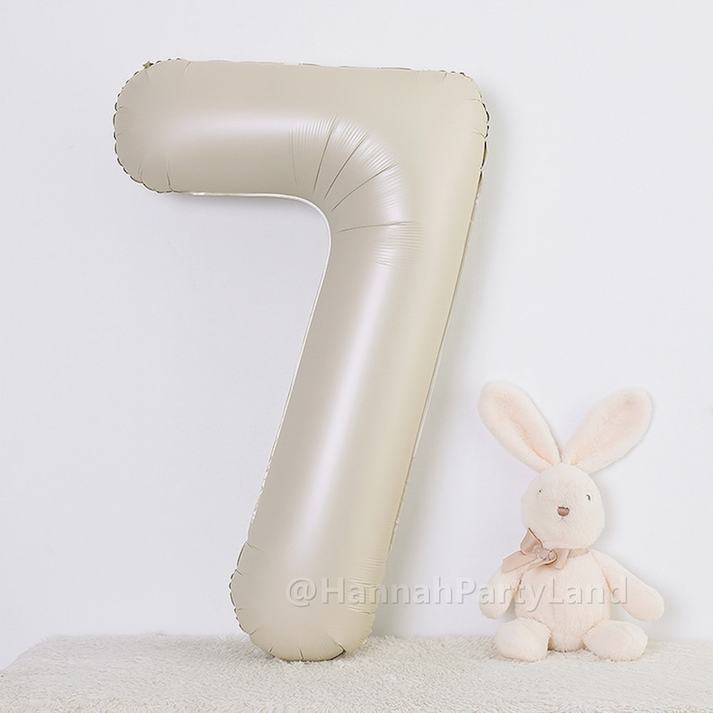 May include: A large, beige, foil number seven balloon. The balloon is standing upright on a white, fluffy surface. A small, white, plush bunny is sitting in front of the balloon.