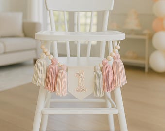 May include: A white wooden high chair decorated with a banner. The banner features a cream-coloured pennant with a glittery number one. The banner is adorned with pink and cream tassels and wooden beads. The background includes a sofa, balloons, and a cake.