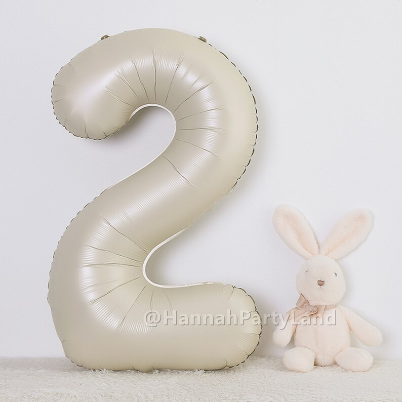May include: A large, beige, foil number two balloon. The balloon is standing upright on a white, fluffy surface. A small, white, plush bunny is sitting to the right of the balloon.