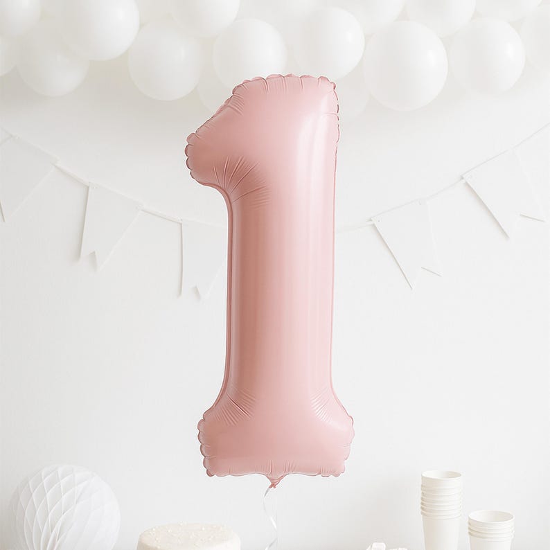 May include: A large, pink number one balloon, perfect for a first birthday. The inflated balloon is set against a white backdrop with white balloons and a banner. The balloon is approximately 76 cm tall.
