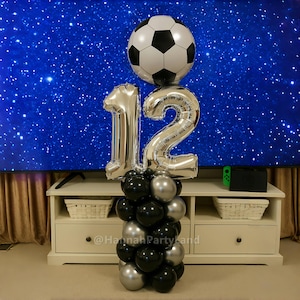May include: A silver number 12 balloon with a black and white football balloon on top. The balloons are on a black and silver balloon column. The balloons are in front of a white entertainment unit with a television and a green video game console.