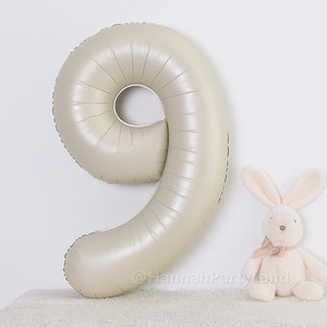 May include: A large, beige, metallic balloon shaped like the number nine. The balloon is inflated and has a shiny finish. It is sitting on a white surface.