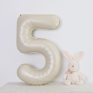 May include: A large, beige, foil number five balloon. The balloon is partially inflated and has a slight sheen. A small, white, plush bunny is sitting to the right of the balloon. The text "@HannahPartyLand" is visible at the bottom of the image.