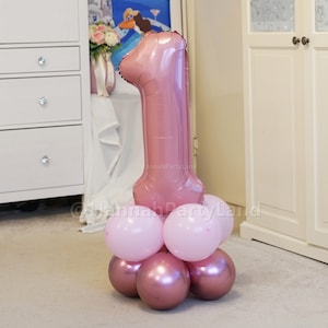May include: A large rose gold number one balloon with a cluster of smaller pink and rose gold balloons at the base.