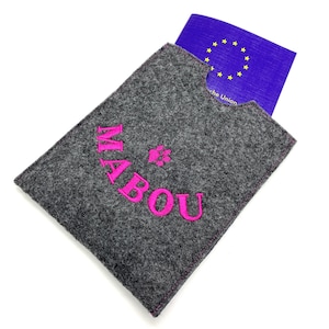 Può includere: Portapassaporto in feltro grigio con ricamo rosa che dice "MABOU" e un'impronta di zampa. Il portapassaporto ha una tasca per un passaporto e il logo dell'Unione Europea è visibile.