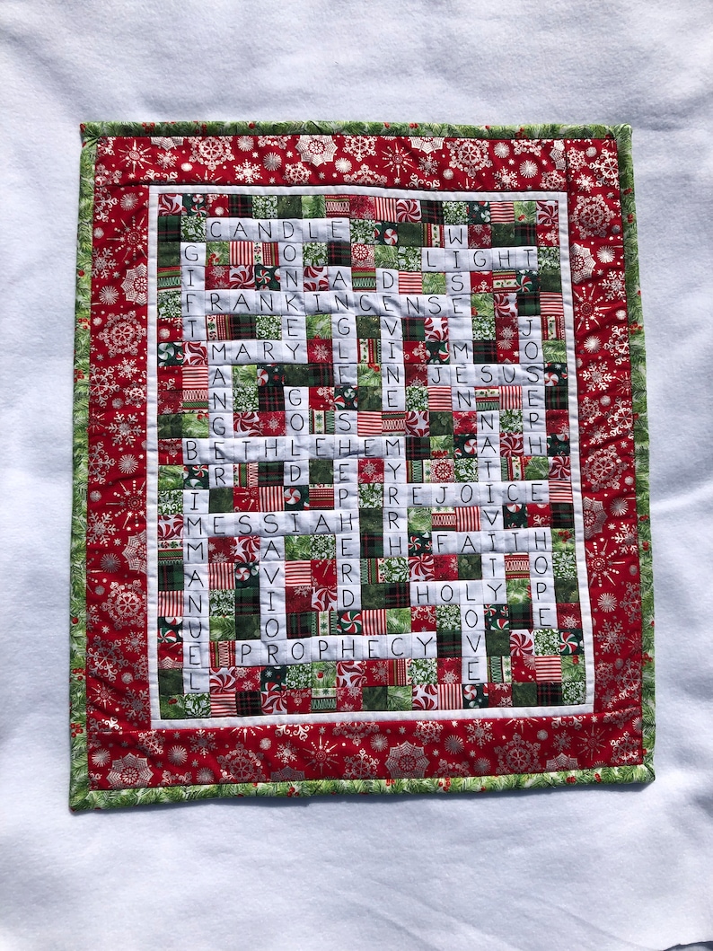 May include: A red and green Christmas-themed crossword puzzle quilt with a white background. The quilt features a variety of Christmas-themed fabrics, including snowflakes, candy canes, and holly. The crossword puzzle features words related to Christmas, such as "candle", "light", "frankincense", "mary", "jesus", "bethlehem", "messiah", "rejoice", "faith", "holy", "love", and "prophecy".