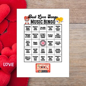 May include: A printed "Best Love Songs Music Bingo" game card with song titles and years. The card features a red cassette tape graphic and heart accents. The background includes red heart decorations and a wooden surface.