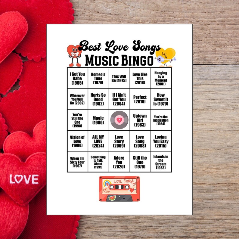 May include: A printed "Best Love Songs Music Bingo" game card with song titles and years. The card features a red cassette tape graphic and heart accents. The background includes red heart decorations and a wooden surface.