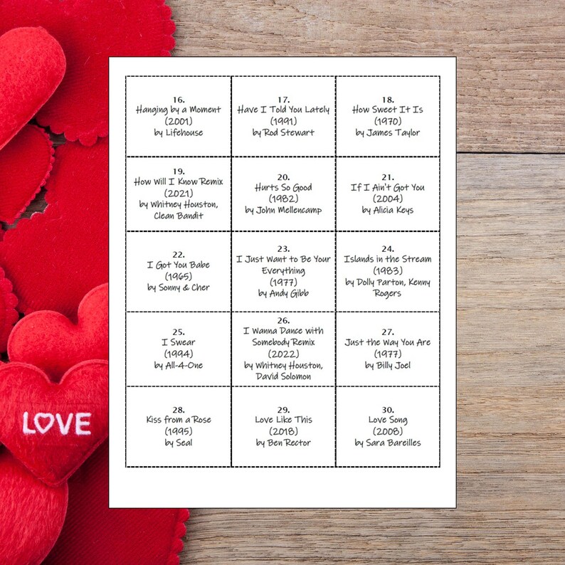 May include: A white card with a grid of song titles and artists, including "Hanging by a Moment" and "How Sweet It Is." Red heart-shaped decorations with the word "LOVE" are in the foreground.