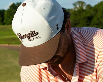 Könnte beinhalten: Eine weiß-braune Baseballkappe mit dem Text "Georgia GOLF CO." in brauner Schrift. Die Kappe hat einen gebogenen Schirm und einen kleinen braunen Knopf oben. Die Person trägt ein gestreiftes orange-weißes Poloshirt.