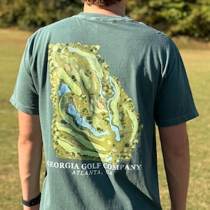 May include: A green t-shirt with a graphic of a golf course in the shape of the state of Georgia. The text "GEORGIA GOLF COMPANY ATLANTA, GA" is printed below the graphic.