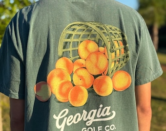 Op de afbeelding: Groen t-shirt met een afbeelding van een mand vol perziken. De tekst "Georgia" is in een boog boven "GOLF CO." in het wit geplaatst. De perziken zijn oranje en geel, en de mand is groen.