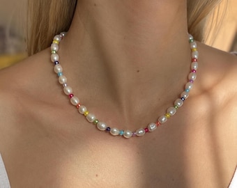 May include: A necklace featuring a repeating pattern of white oval-shaped pearls interspersed with small, round, multi-colored beads in shades of red, orange, yellow, green, blue, and purple, arranged in a single strand around the neck.