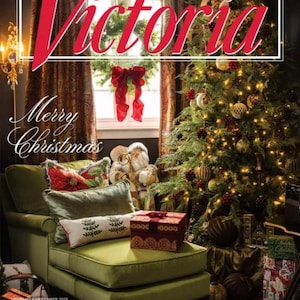 Puede incluir: La portada de la revista "Victoria" presenta una escena navideña festiva. Un árbol de Navidad decorado con luces y adornos se encuentra cerca de una chaise longue verde con cojines. El texto "Merry Christmas" es visible, junto con el título de la revista.