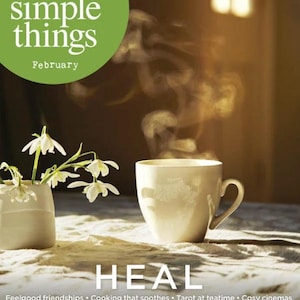 Può includere: Una copertina di rivista con il titolo "the simple things" e il mese "February". Una tazza bianca con vapore che sale è su un tavolo. La parola "HEAL" è sotto.