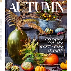 Op de afbeelding: De cover van Country Living magazine toont het woord "AUTUMN" in grote witte letters. De afbeelding toont een seizoensgebonden opstelling met pompoenen, kalebassen en herfstbladeren. Een grote groene fles en een metalen emmer gevuld met planten zijn ook zichtbaar.
