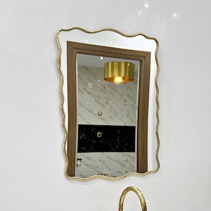 May include: A white marble oval sink with a gold faucet and a gold framed mirror with a wavy edge. The mirror reflects a gold light fixture.