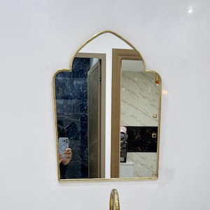 May include: A white marble oval sink with a gold faucet and a gold framed arched mirror. The mirror reflects a bathroom with a tiled floor and a wooden door.