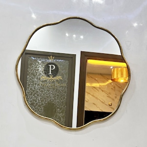 Może przedstawiać: Złote, ozdobne lustro z falistymi krawędziami wisi na białej ścianie nad złotą baterią. Lustro odbija znak z tekstem "P Pristine Brass".