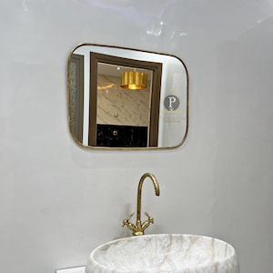 May include: A white marble oval sink with a gold faucet and a gold framed rectangular mirror above it. The mirror reflects a room with a gold light fixture.