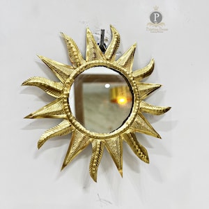 Puede incluir: Un espejo de sol dorado con un espejo redondo en el centro. El espejo está rodeado por un marco decorativo con un diseño de sol. El espejo está colgado en una pared blanca.