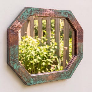 May include: An octagonal wall mirror featuring a hammered copper frame with a weathered patina finish. The metal surface displays a blend of warm metallic copper tones and oxidised teal green accents, creating a textured, aged appearance.