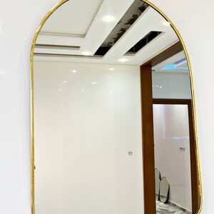 Puede incluir: Un espejo arqueado con marco dorado refleja un techo blanco con iluminaci&oacute;n empotrada y una puerta que conduce a otra habitaci&oacute;n.