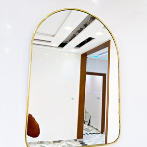 Puede incluir: Un espejo arqueado con marco dorado que refleja una habitaci&oacute;n blanca con una puerta de madera y suelo de baldosas.