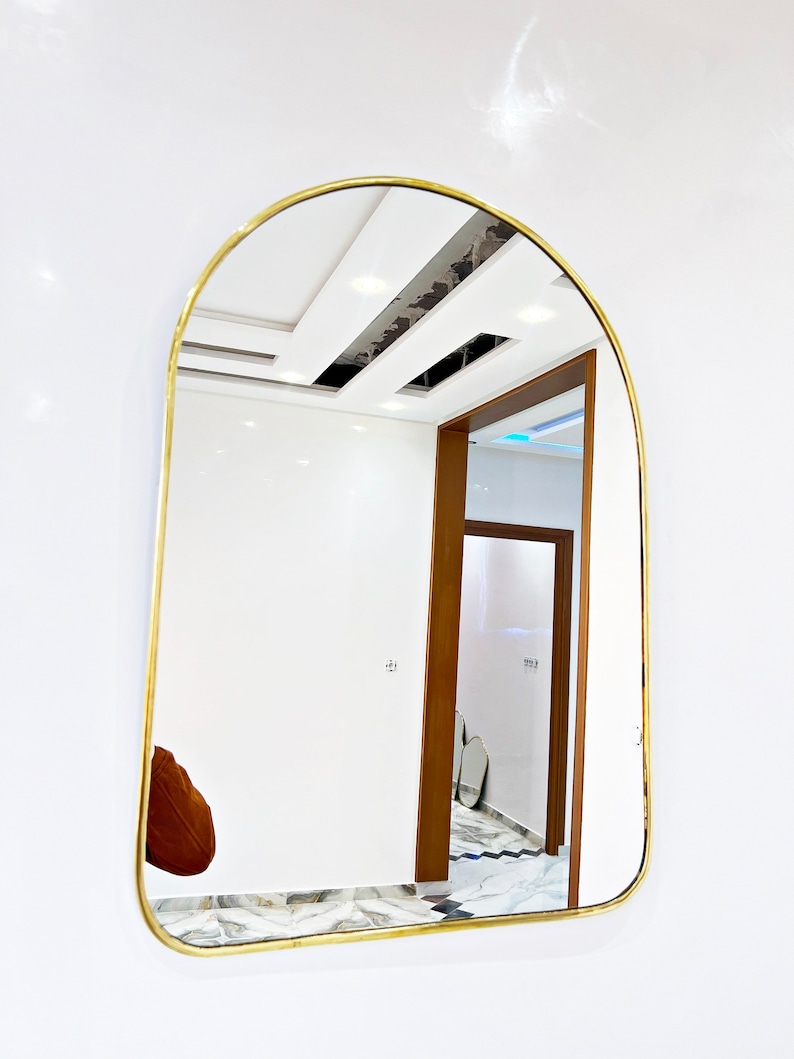 Puede incluir: Un espejo arqueado con marco dorado que refleja una habitaci&oacute;n blanca con una puerta de madera y suelo de baldosas.