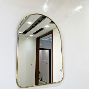 Puede incluir: Un espejo arqueado con marco dorado que refleja un pasillo con techo blanco y una puerta de madera.