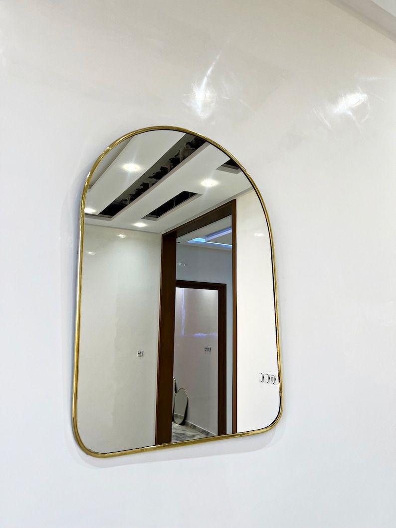 Puede incluir: Un espejo arqueado con marco dorado que refleja un pasillo con techo blanco y una puerta de madera.