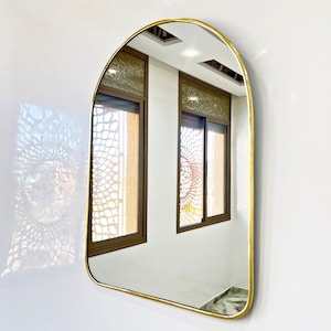 Puede incluir: Un espejo arqueado con marco dorado que refleja dos ventanas con vidrio estampado. El espejo est&aacute; colgado en una pared blanca.