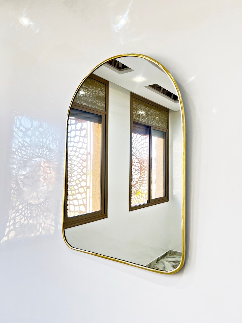 Puede incluir: Un espejo arqueado con marco dorado que refleja dos ventanas con vidrio estampado. El espejo est&aacute; colgado en una pared blanca.