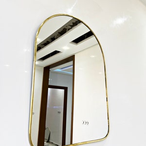 Puede incluir: Un espejo arqueado con marco dorado, parte superior redondeada y superficie lisa y reflectante. El espejo est&aacute; montado en una pared blanca y refleja un pasillo con una puerta de madera.